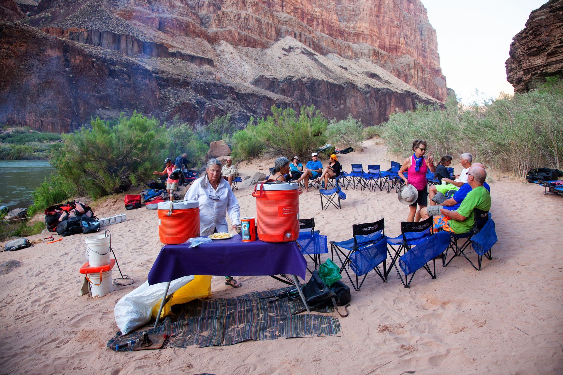People at a riverside campsite, setting up chairs and a table. Red and blue coolers, canyon backdrop.