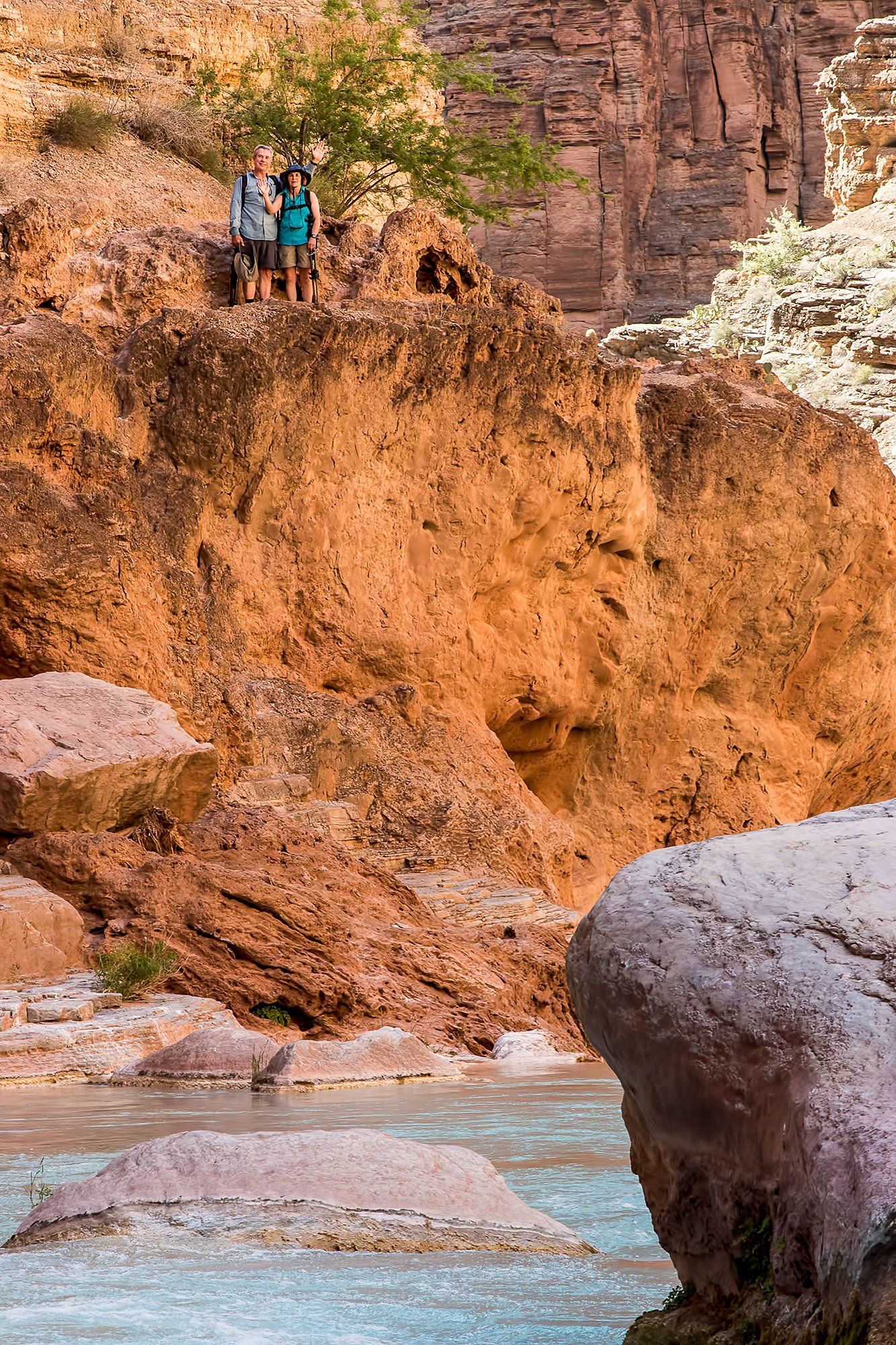 Two people stand on a cliff edge, overlooking a turquoise river flowing through a canyon.