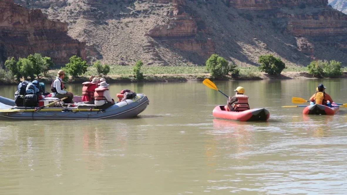 Rafting boats on a river near rocky cliffs; two kayakers paddle alongside.