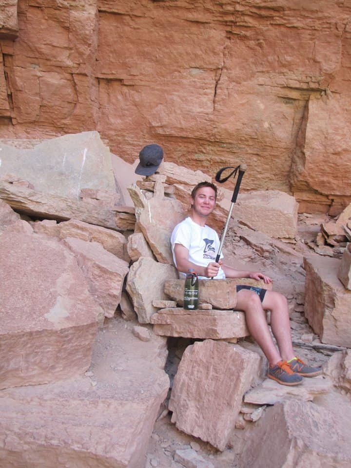 Man sitting on rocks in a red rock canyon, holding a hiking pole and beverage. Black hat on rock.