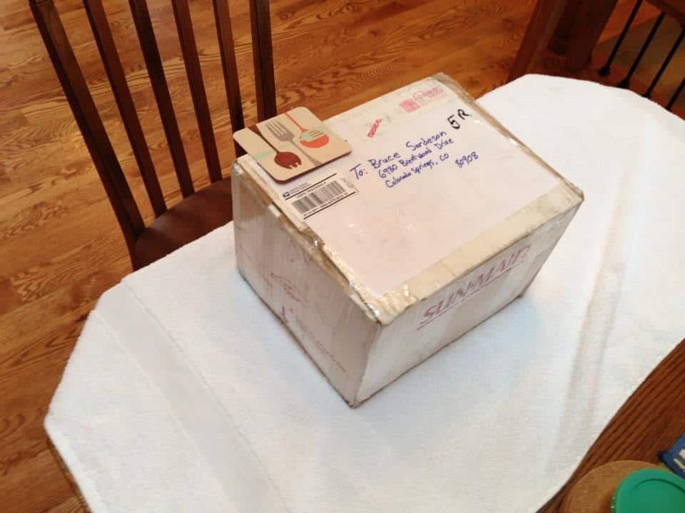 Cardboard box on a white tablecloth, with a coaster and address label visible. A wooden chair is nearby.