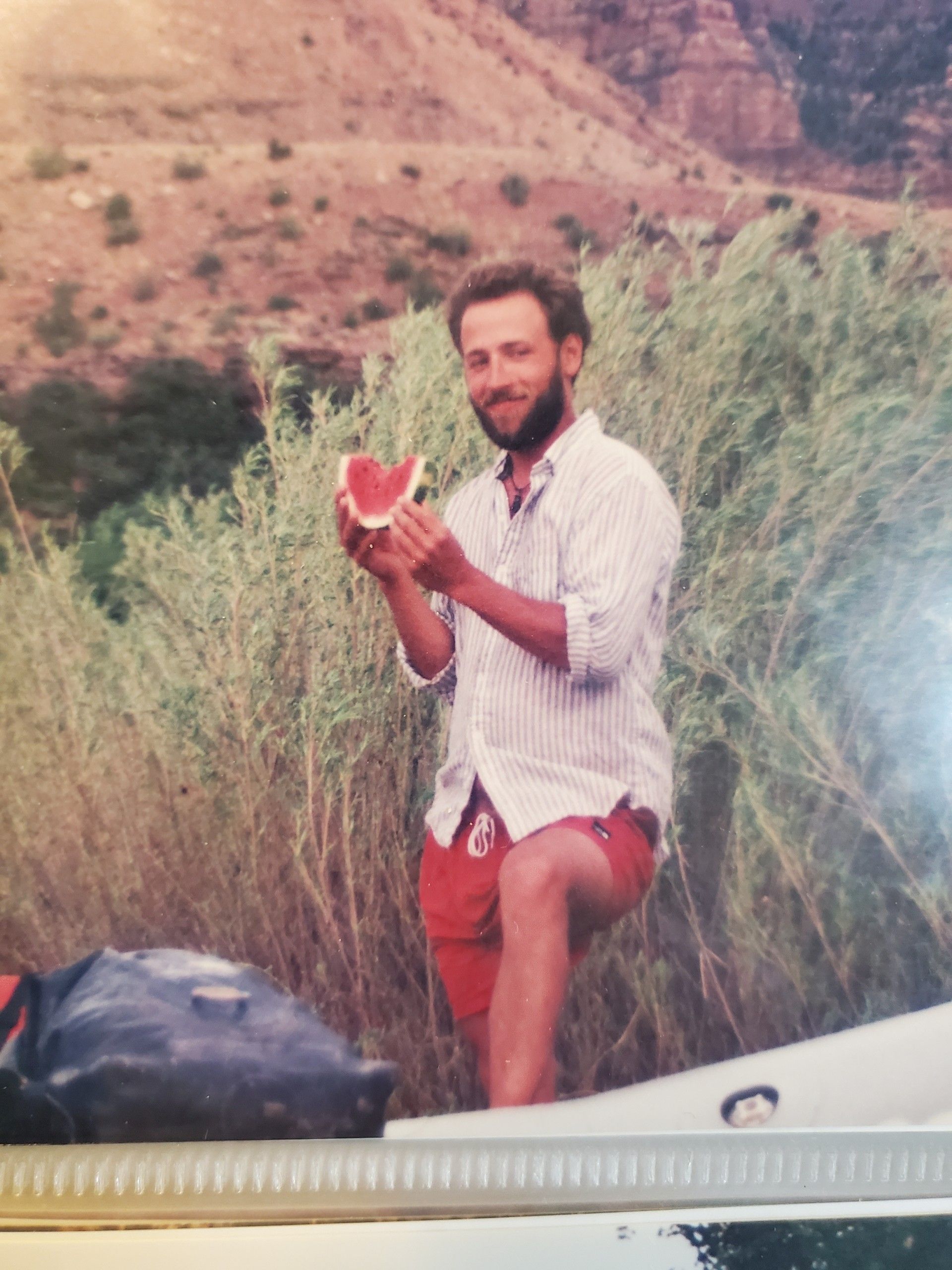 Man eating watermelon outdoors, red shorts, holding a slice, arid background.