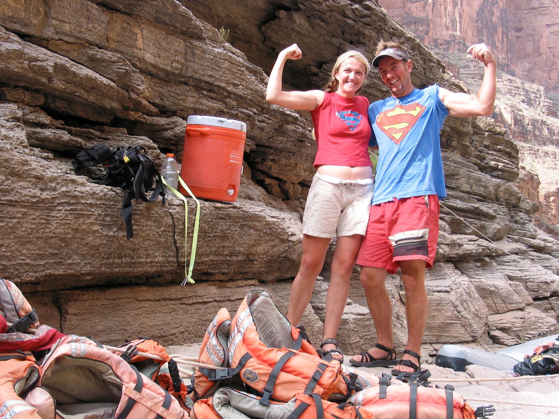 Couple flexing arms, standing near rocks, wearing casual clothes and sandals, by life jackets.