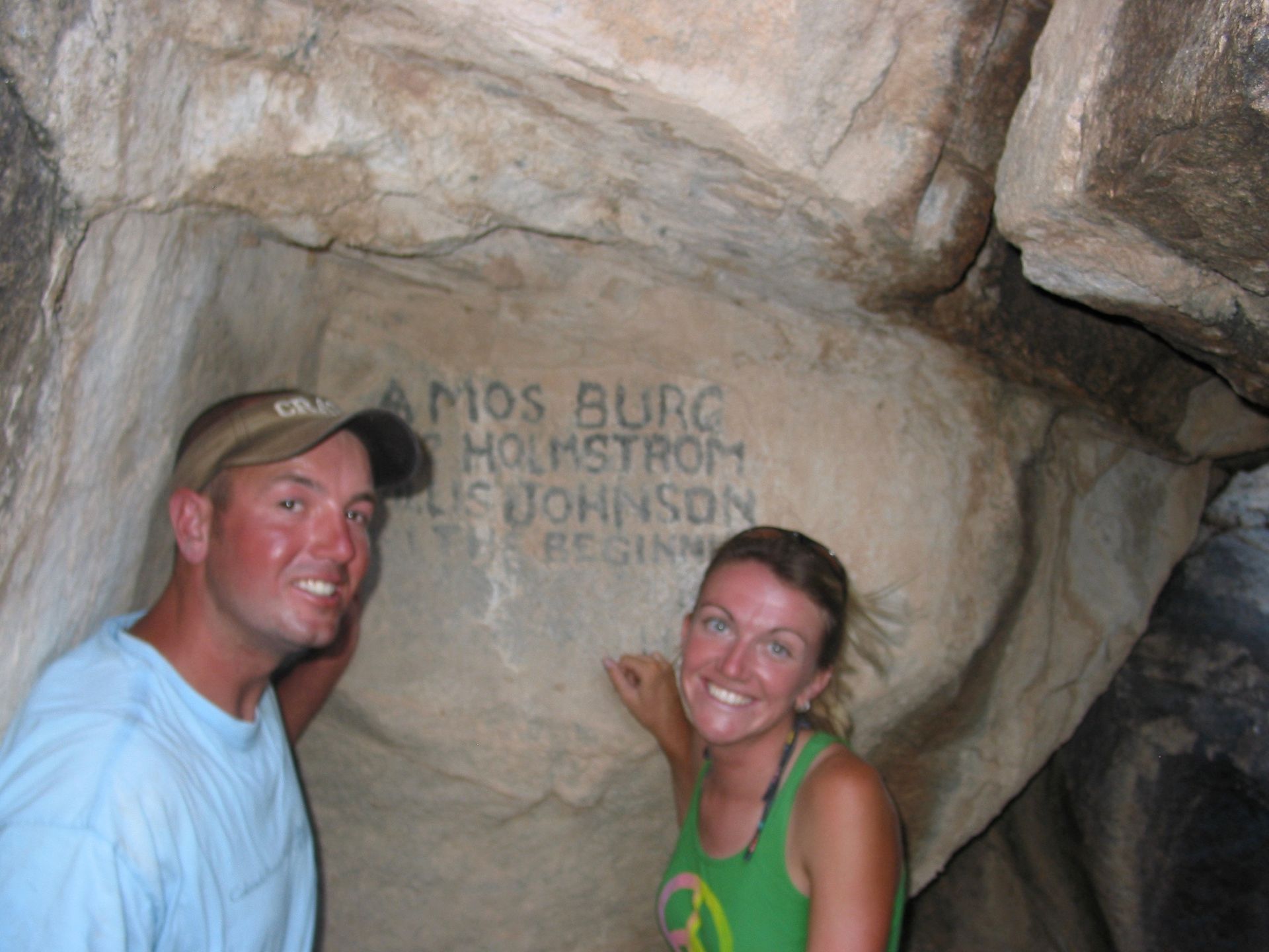 Man and woman smiling by carved names in a rock cave: Amos Burg, Holstrom, and Ellis Johnson.