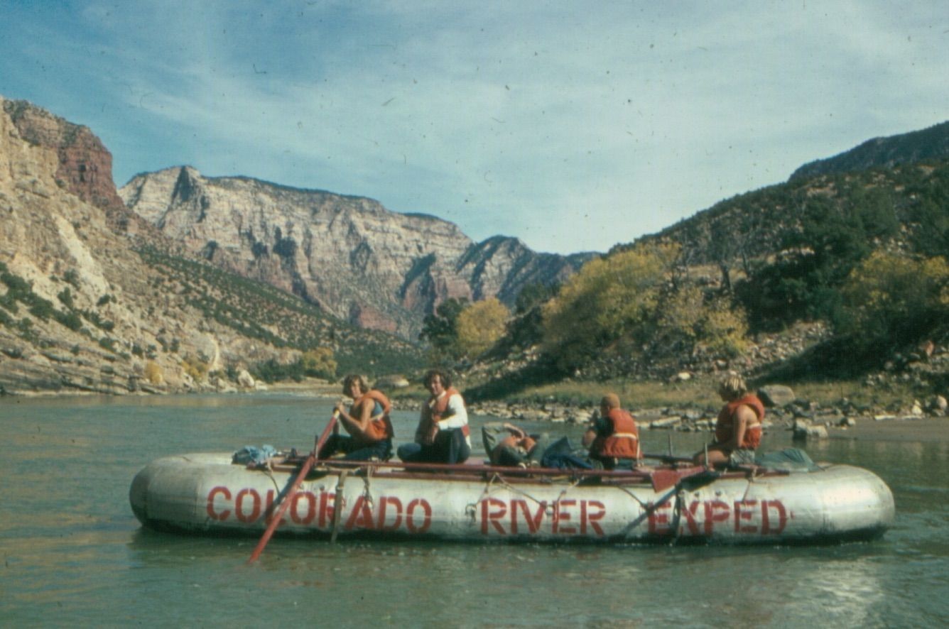 Rafting on Colorado River. People in orange life vests on inflatable raft, with rocky canyon landscape.
