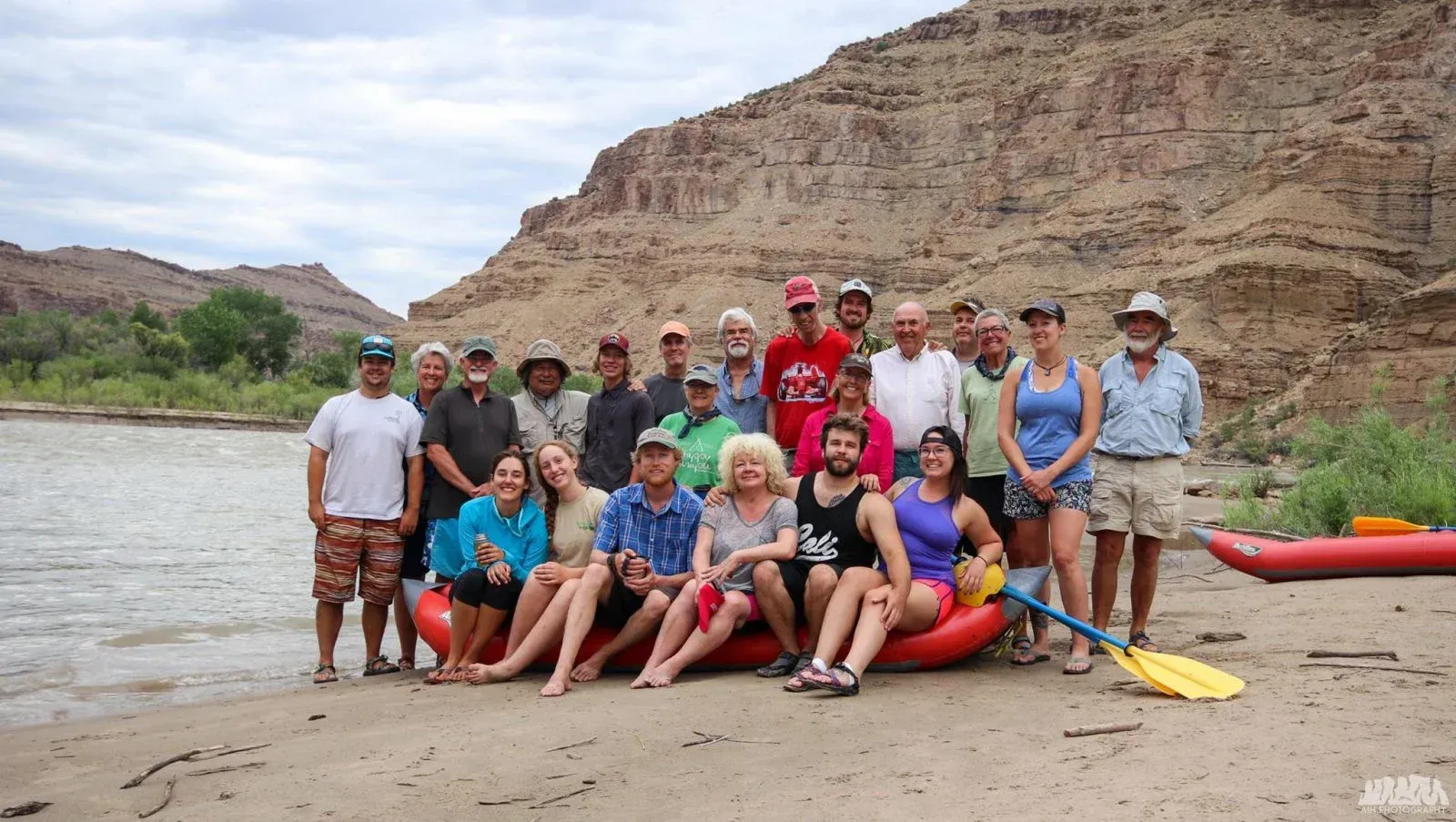 Group poses on a riverbank with raft and backdrop of cliffs.