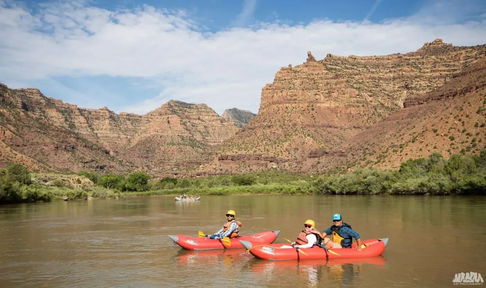 People in red rafts paddling on a river, with red rock mountains in the background under a blue sky.