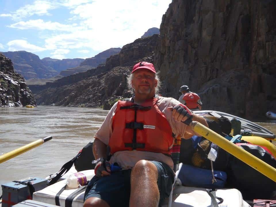 Man in life vest rows raft on a river, canyon walls in the background. Sunny day.