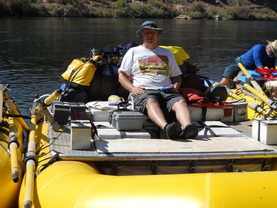 Man seated on a yellow raft, legs crossed. Surrounded by gear and supplies, on a river.