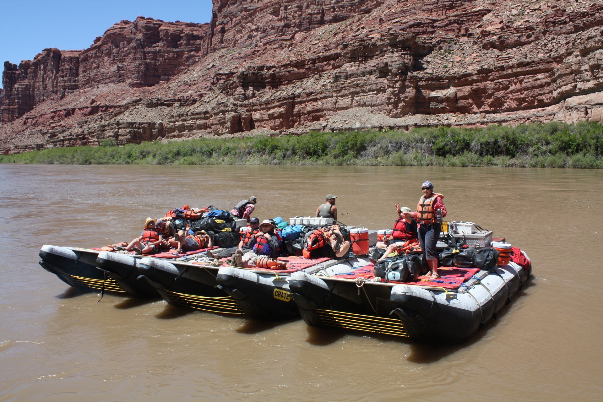 Four rafts on a river with gear and people. Red rock cliffs in the background.