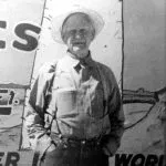 Man in hat and work clothes stands in front of a sign.
