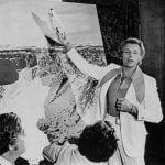 A person in a light suit points at a snowy landscape, lecturing to a seated group.