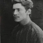 Man in a dark shirt, looking to the side with a mustache, against a dark backdrop.