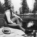 Man with long beard sits by a pond in the mountains. He wears a hat and vest.