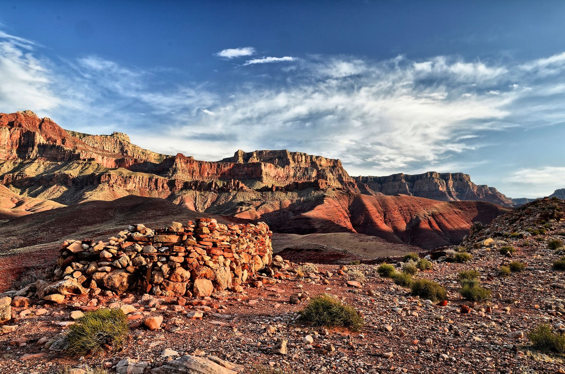 Red rock canyon under a blue sky with streaky clouds; foreground has a rock formation and desert scrub.