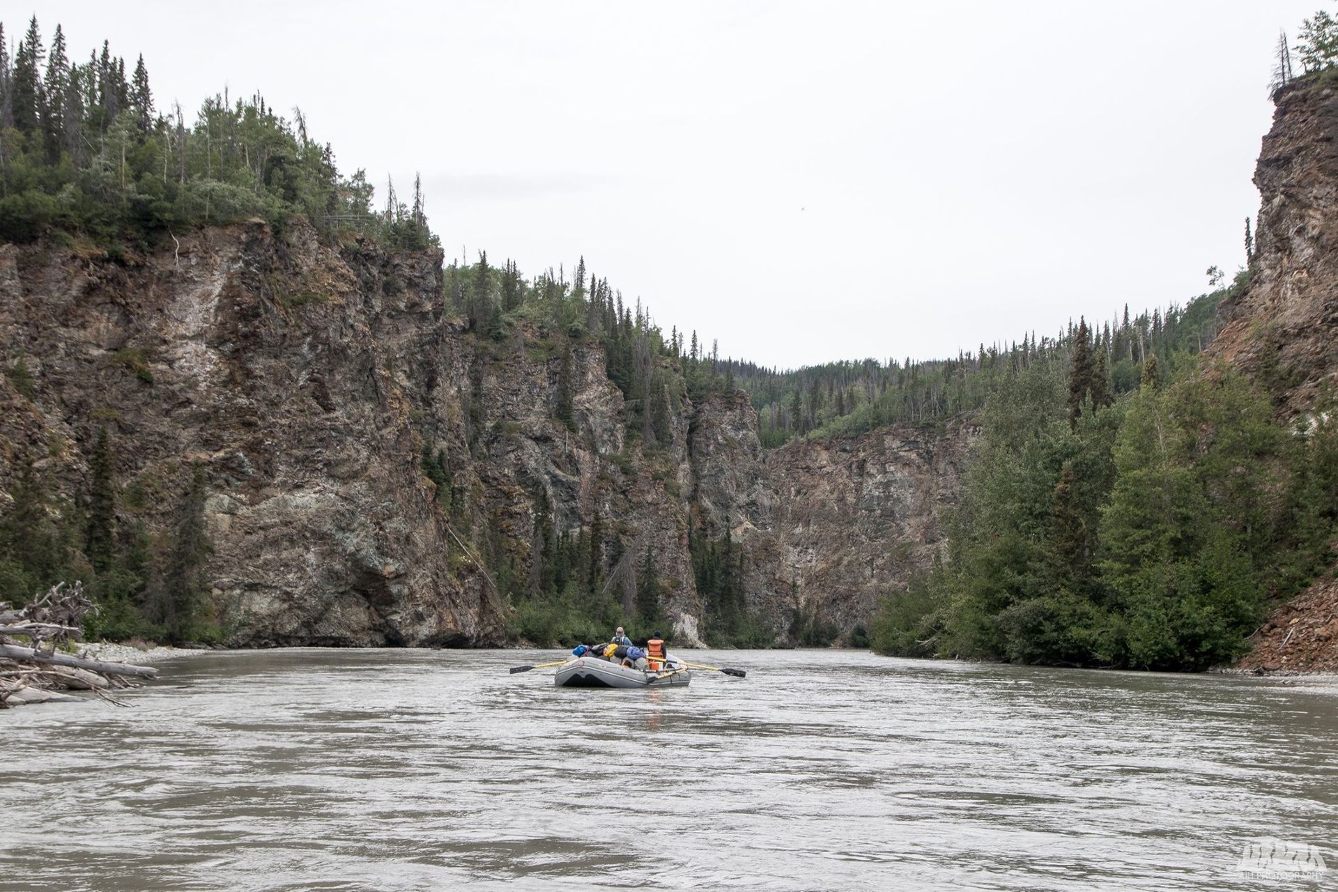 A river flows through a canyon, with two people in a raft. Gray water, rocky cliffs, and green trees.