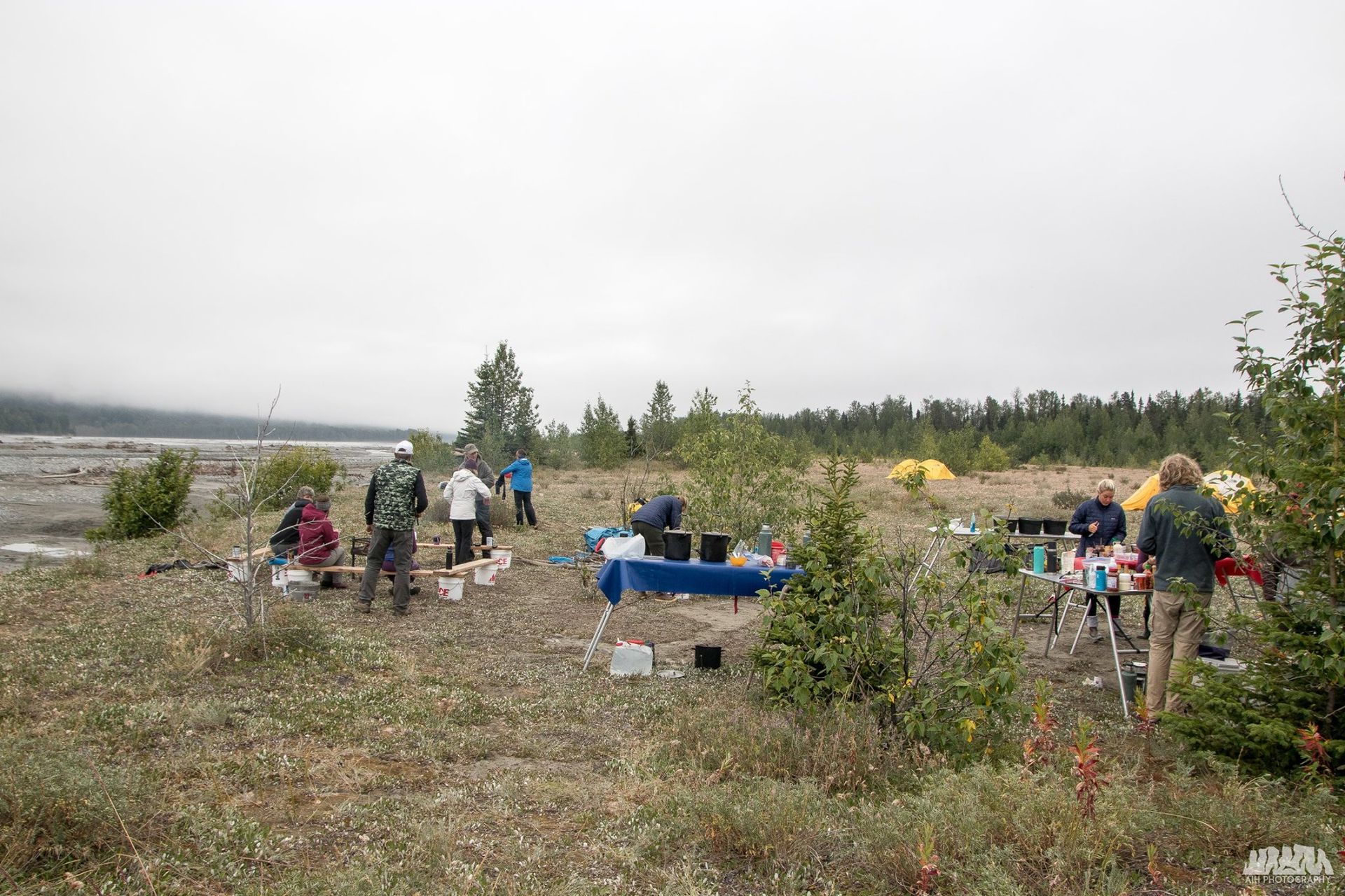 People setting up a camp in a cloudy, grassy area near a river and trees.