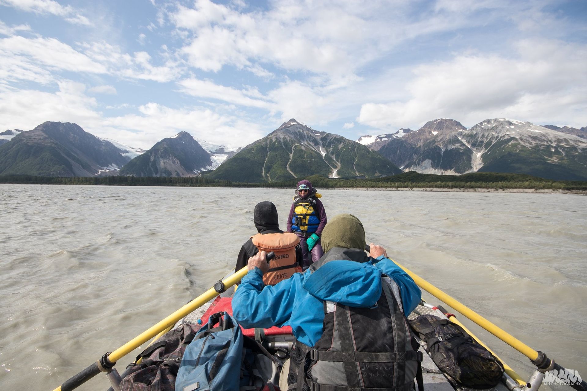 People rowing a raft on a glacial lake, mountains in the background under a cloudy sky.
