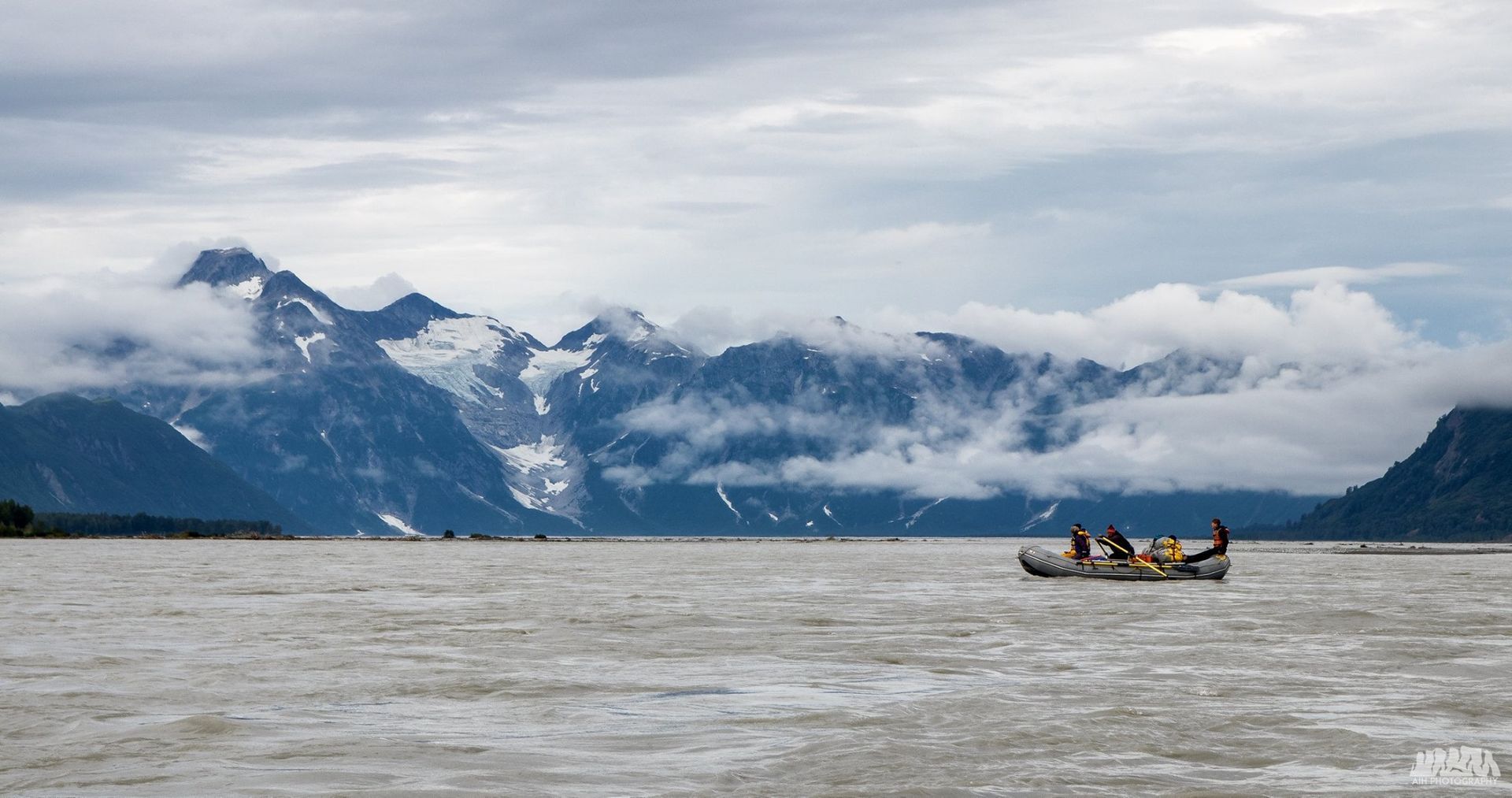 A boat with people navigates a misty lake with mountains in the background. Cloudy sky.