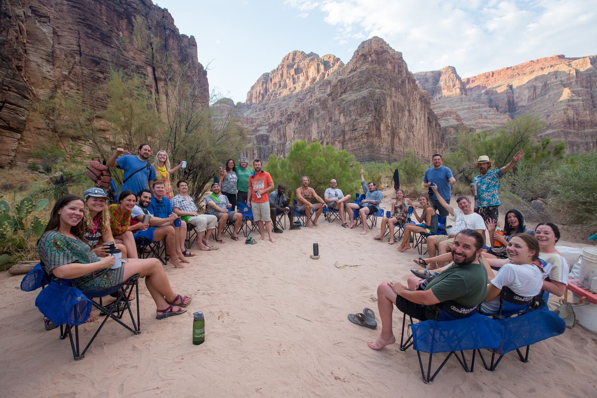 Group of people sitting in a circle on a sandy bank in a canyon. Mountain backdrop, sunny day.
