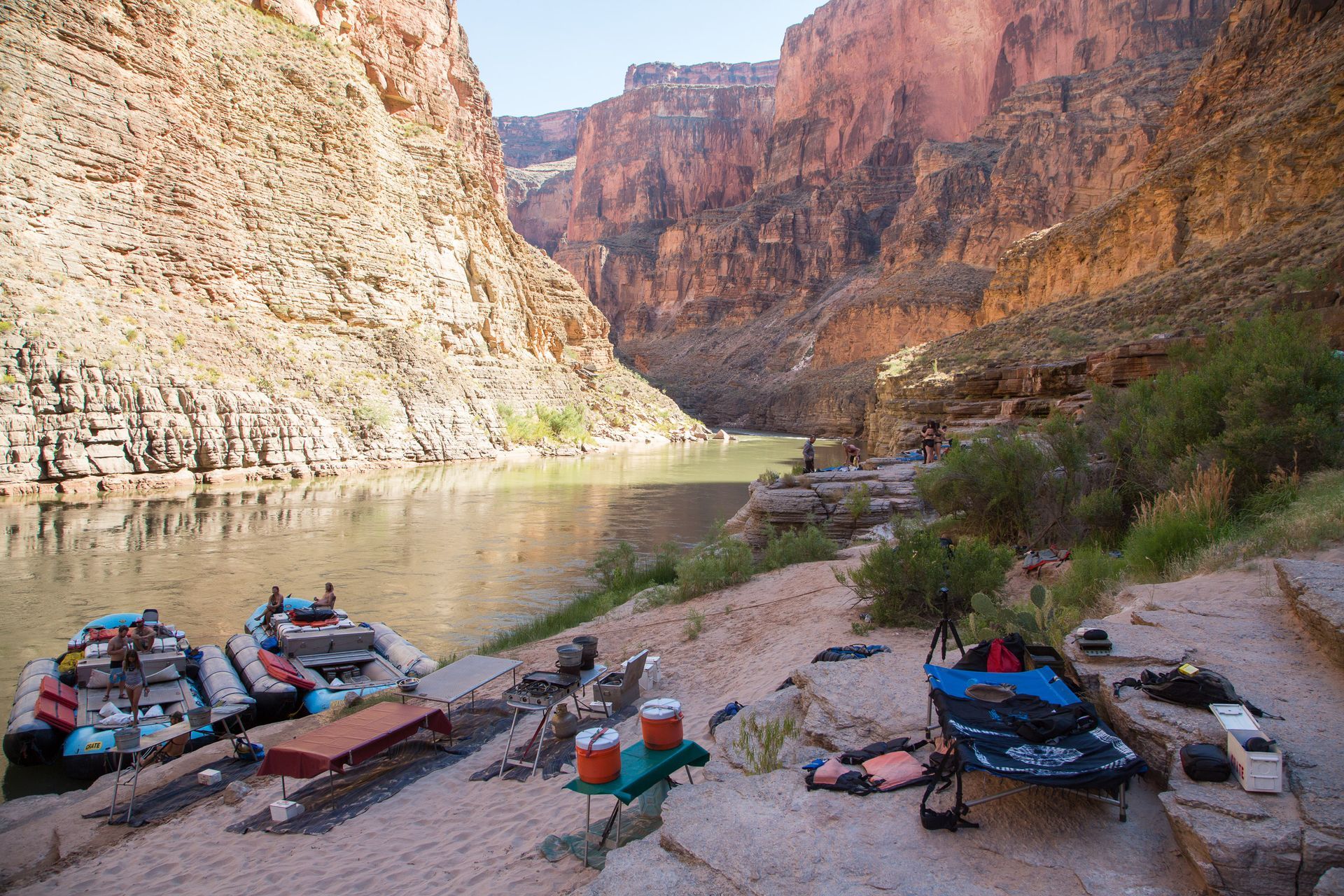 Rafts and gear on a sandy riverbank in a canyon with towering red rock walls, under a sunny sky.