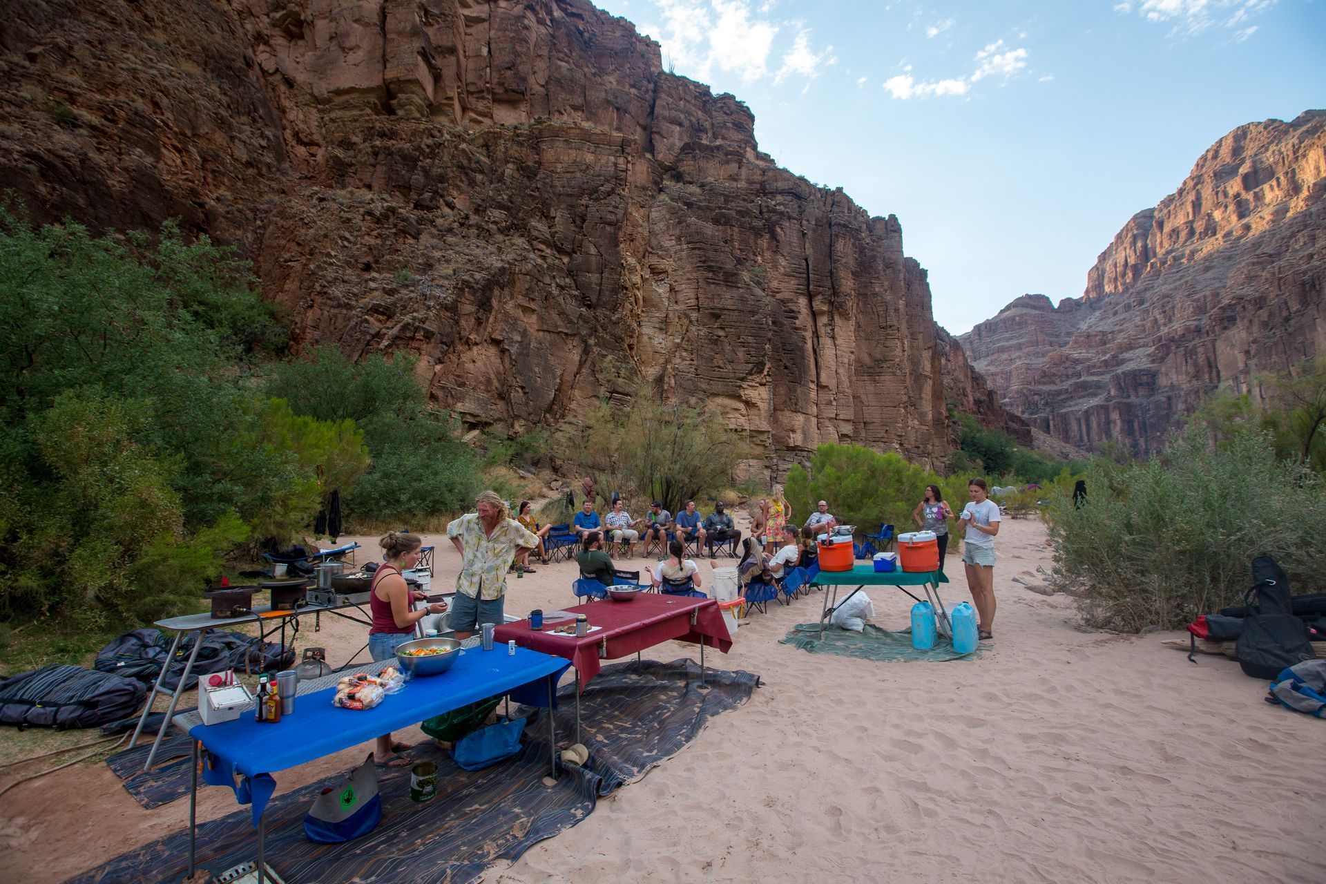 Group camping in a canyon. People set up tables with food and supplies on a sandy beach. Canyon walls surround them.