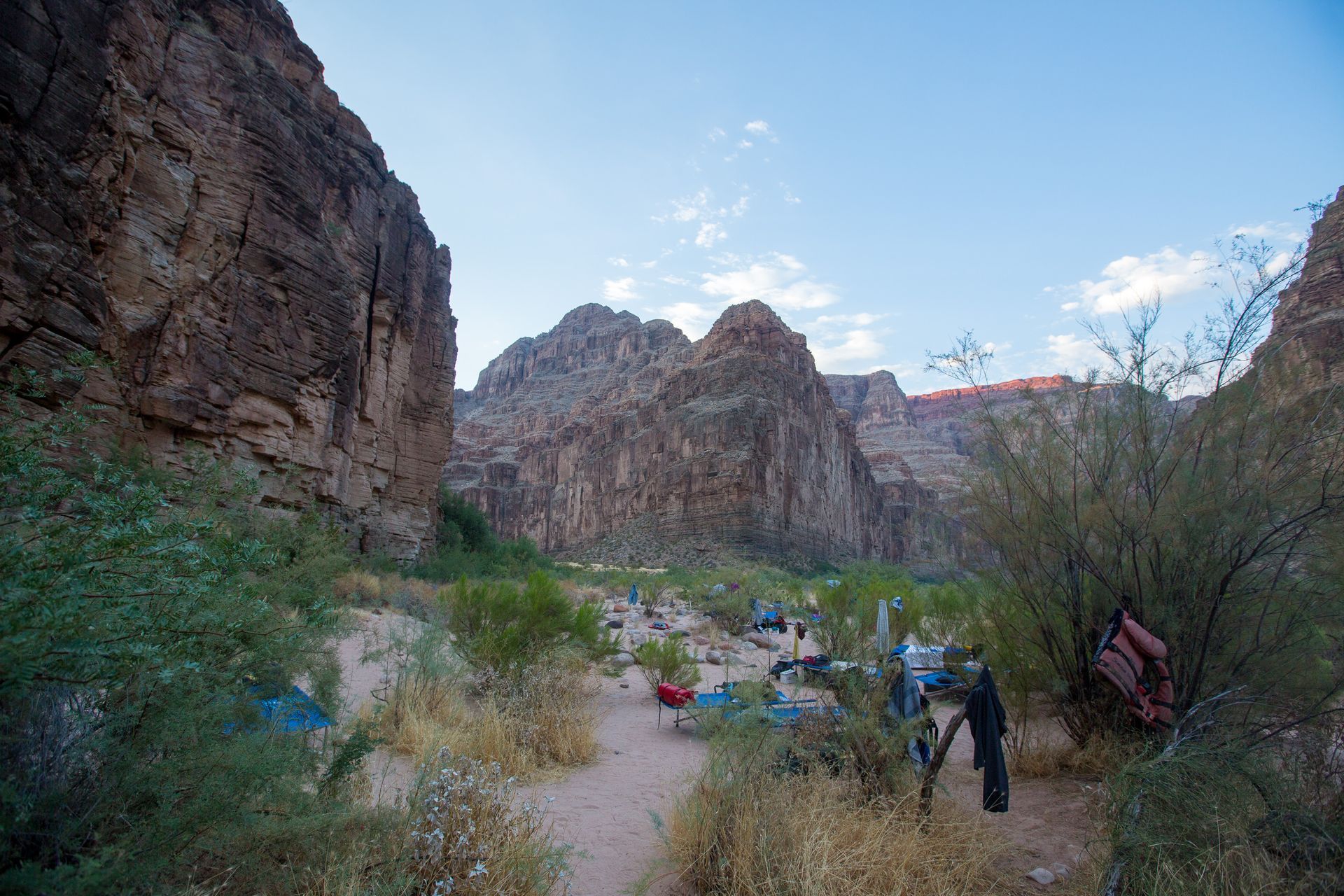 People gather for a meal on a sandy riverbank, with canyon walls in the background. Blue and red tables.