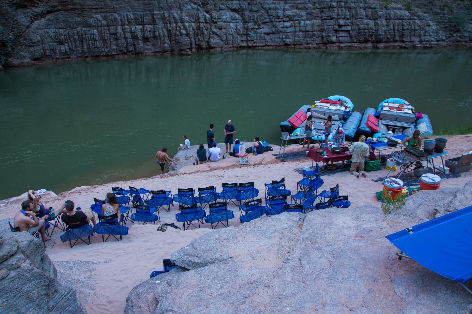 Group of people camp near a river, with rafts, chairs, and gear set up on a rocky shore.