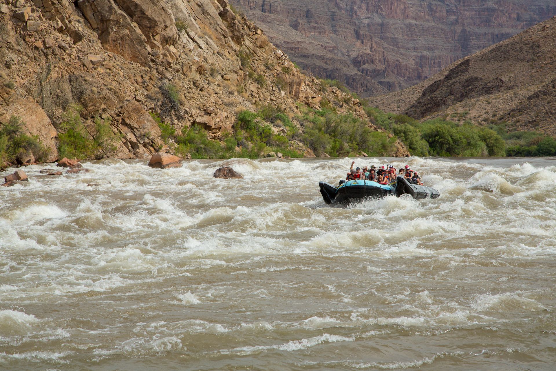 Rafting through whitewater rapids in a canyon with tan rock walls and green bushes.