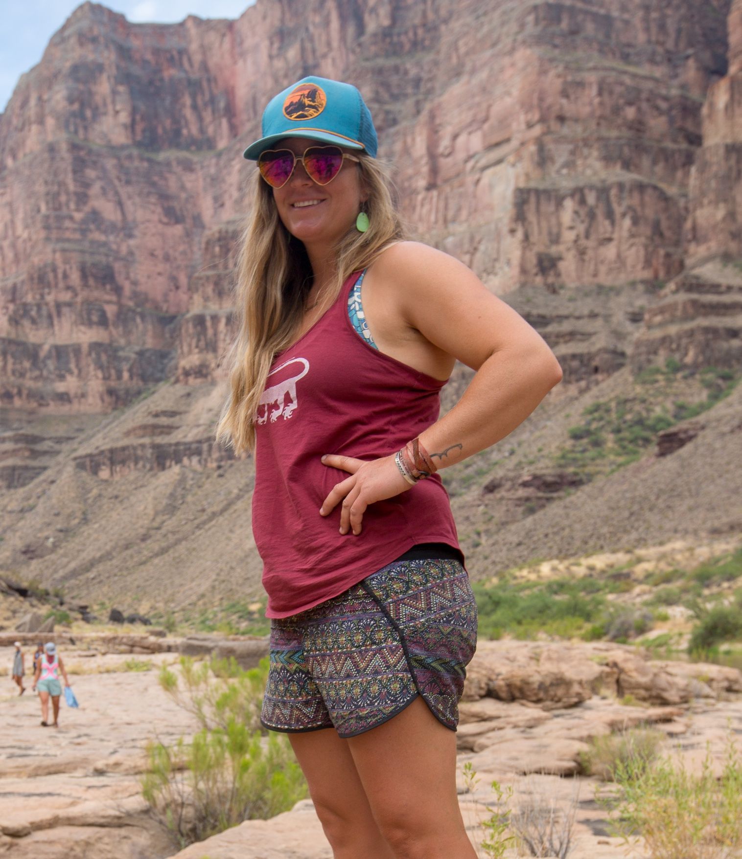Woman in a maroon tank top and patterned shorts, standing outdoors with a mountain backdrop.