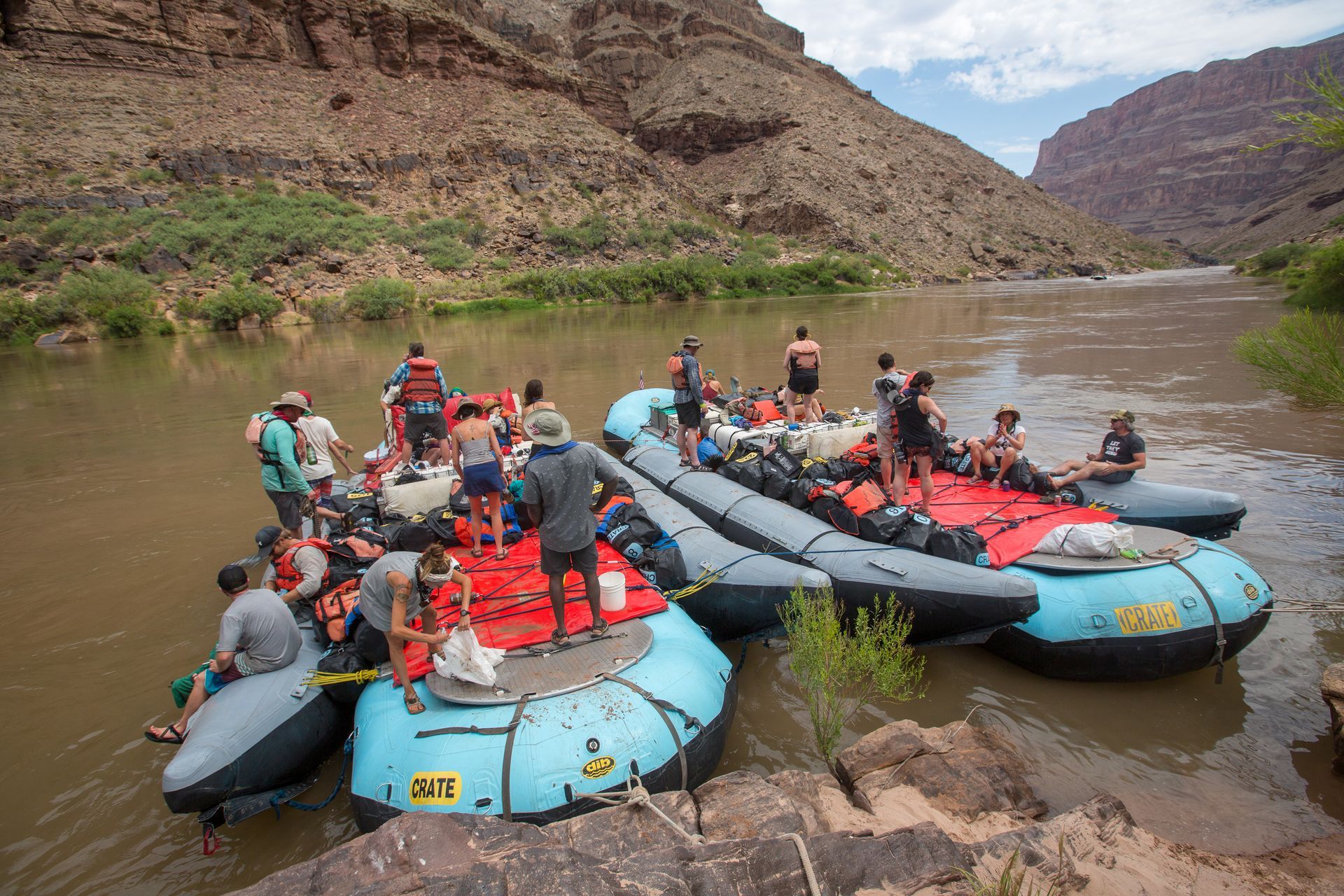 Group of people on rafts on a brown river near canyon walls.
