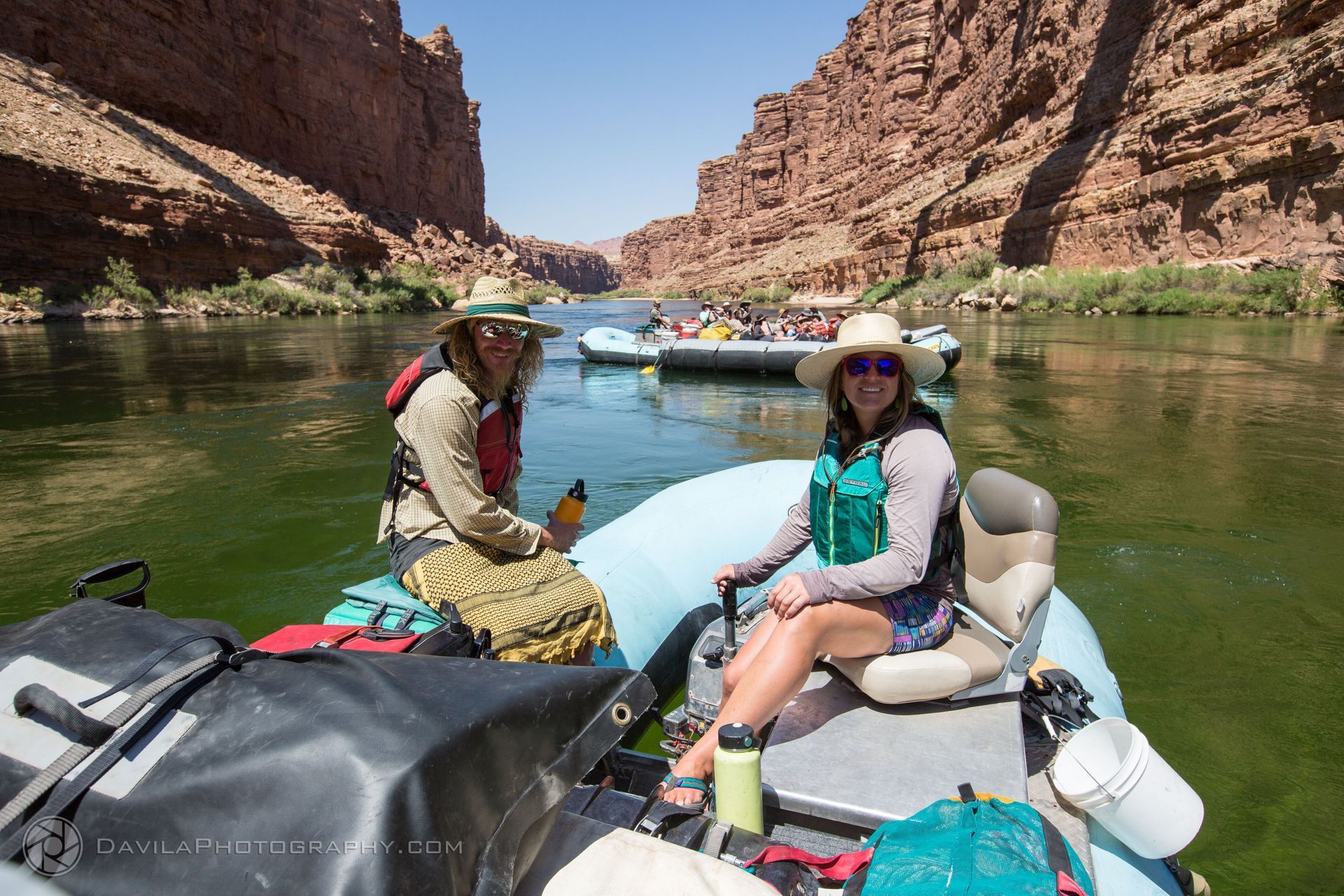 Two people on a raft, floating down a river between red rock canyon walls.