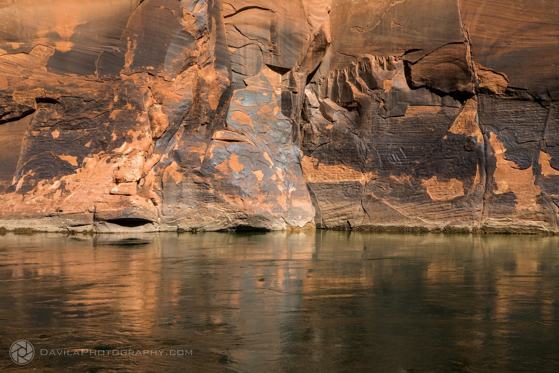 Cliff face of red and black rock above a calm, reflective body of water.