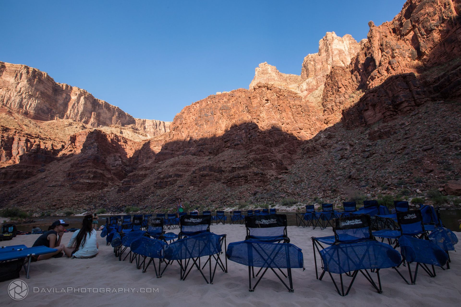 Blue chairs set up for event on a sandy riverbank, towering red rock canyon backdrop.