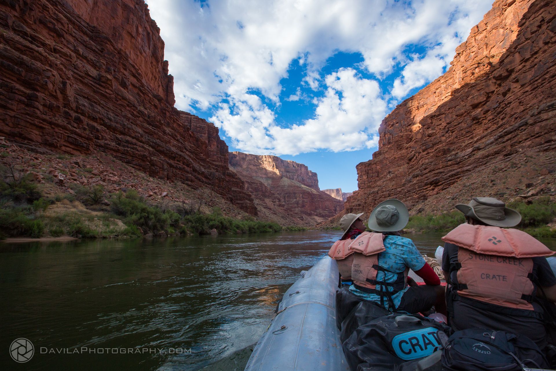Rafting through a red rock canyon. People wear life vests, enjoying the sunny day with blue sky and clouds.