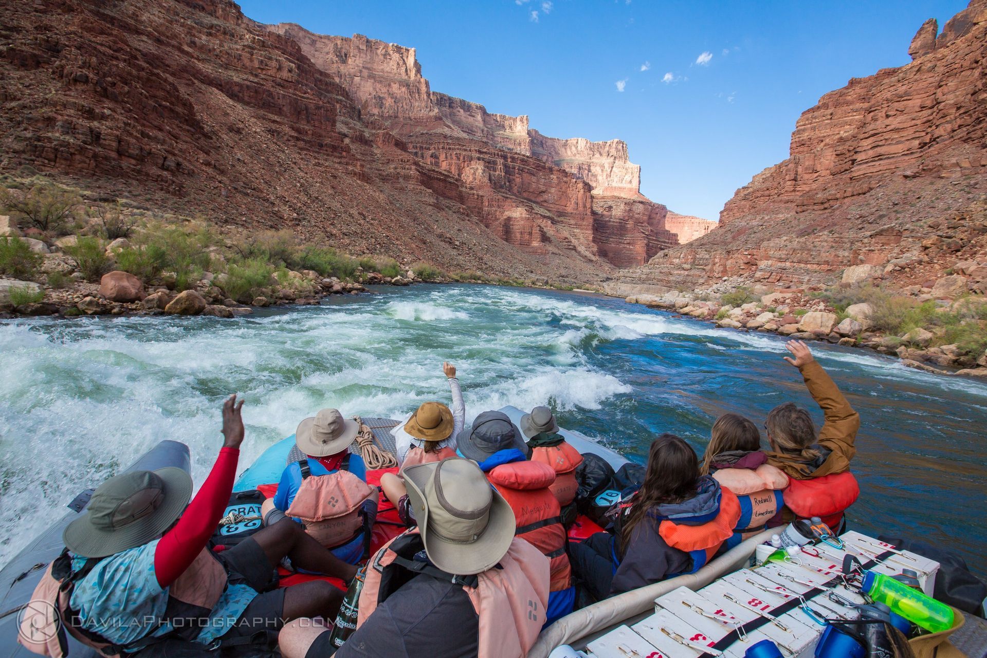 Rafting through a canyon, passengers with raised arms, blue water, red rock walls.