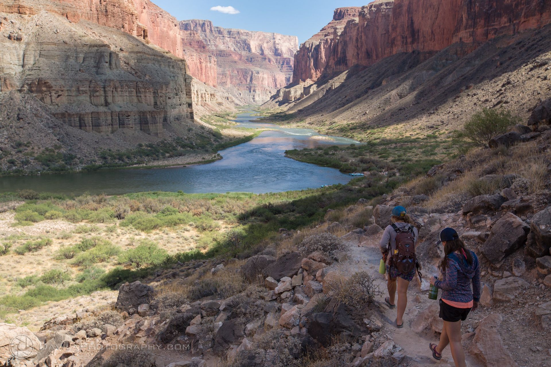 Two hikers on a trail overlooking a river in a canyon, with red rock walls and blue water.