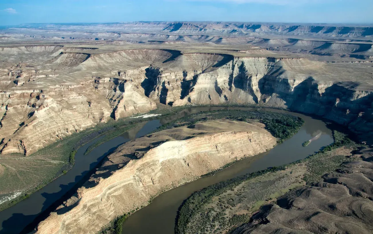 Canyon landscape with a winding river; tan and gray cliffs, blue sky.