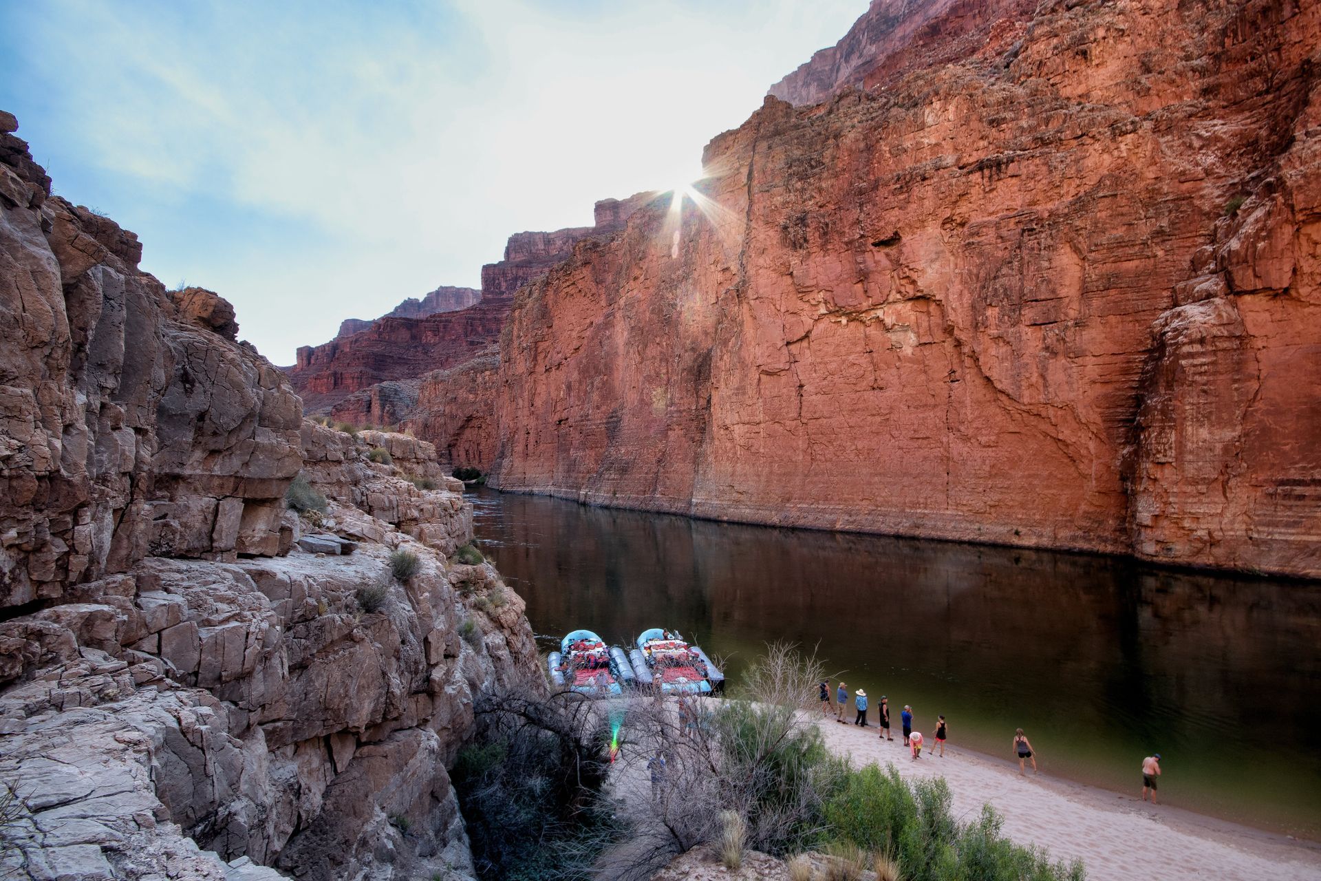 Rafts on a calm river between high, red rock canyon walls. A group of people stand on a sandy shore. Sun shines.