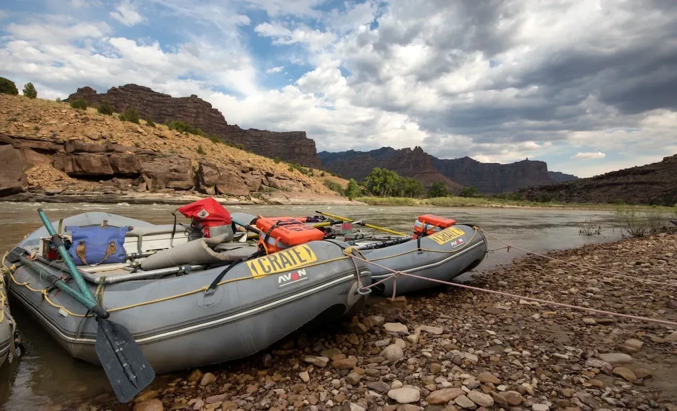 Two gray rafts on a rocky riverbank, brown canyon in the background under a cloudy sky.