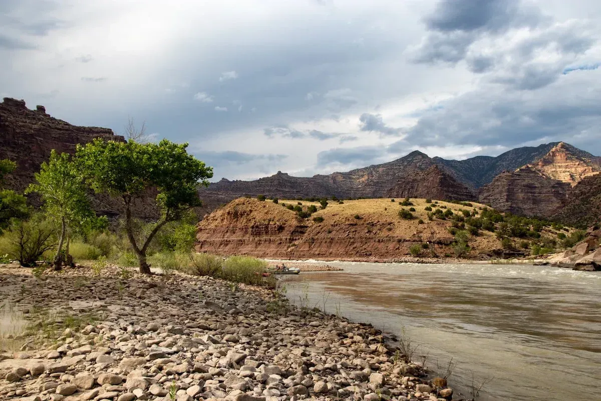 River flowing through arid landscape, mountains in background, cloudy sky.