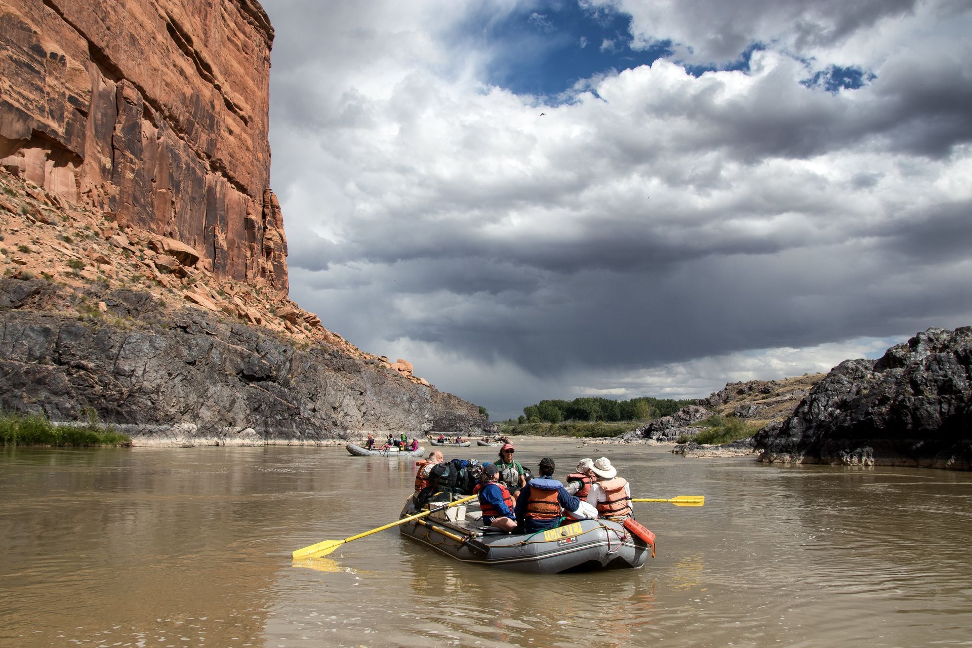 Rafting on a brown river in a canyon. People on raft under cloudy sky, high rock walls on either side.
