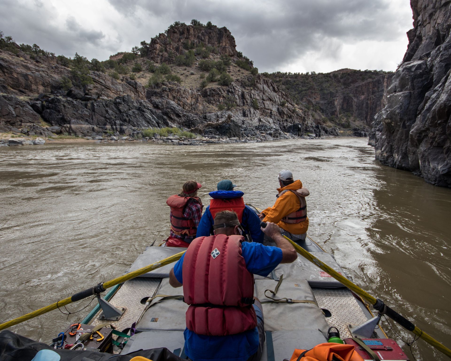 People on a raft navigate a river with canyon walls. Cloudy day.