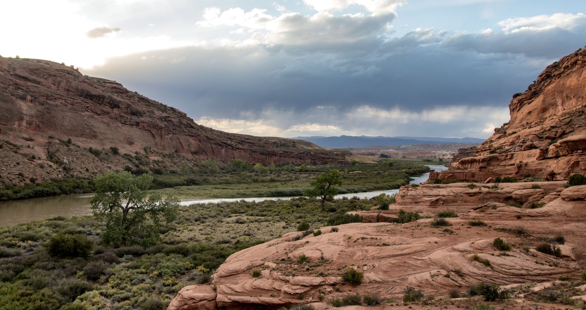 Canyon landscape with river, red rock cliffs, and stormy sky.