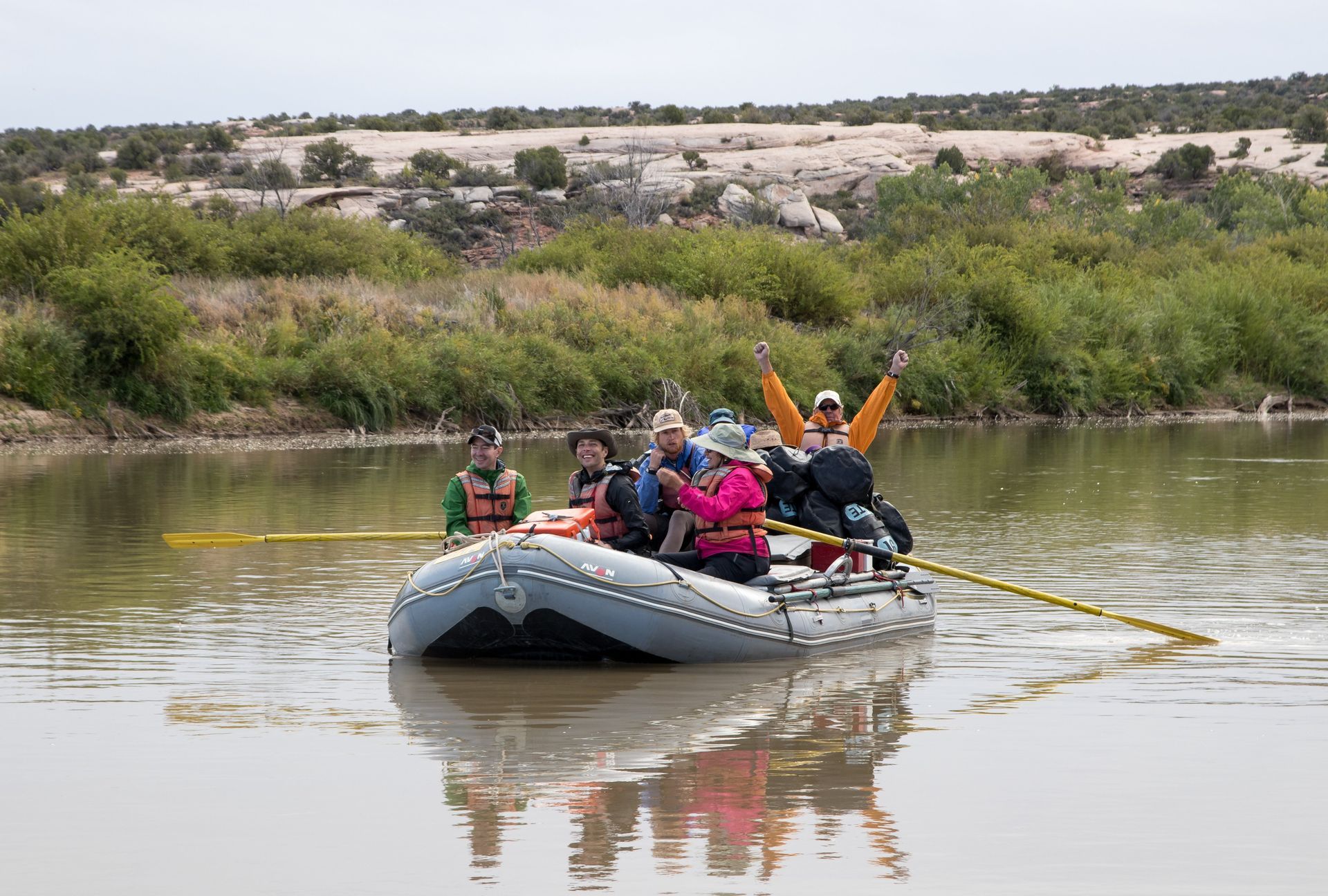 Two people on a raft in a canyon river, wearing life vests and hats.