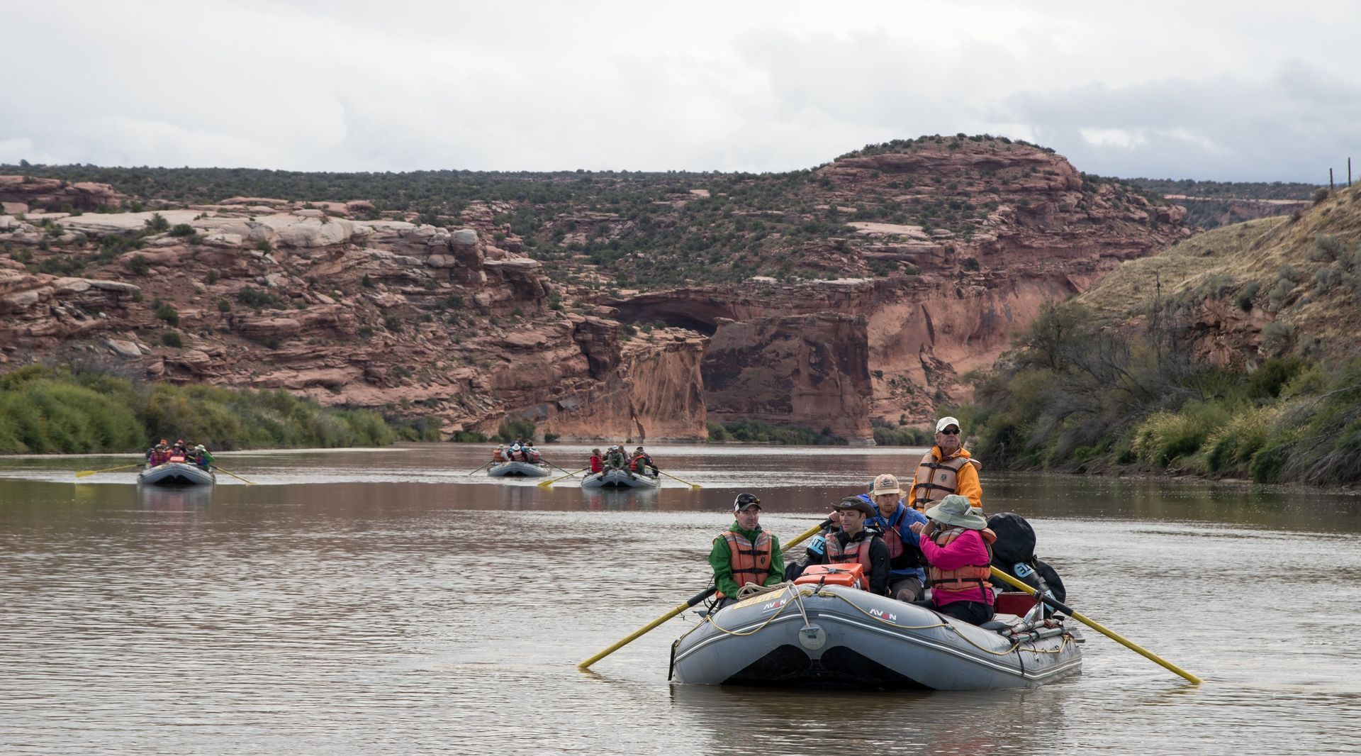 Rafts of people on a river, brown water, red rock canyon in background, cloudy day.
