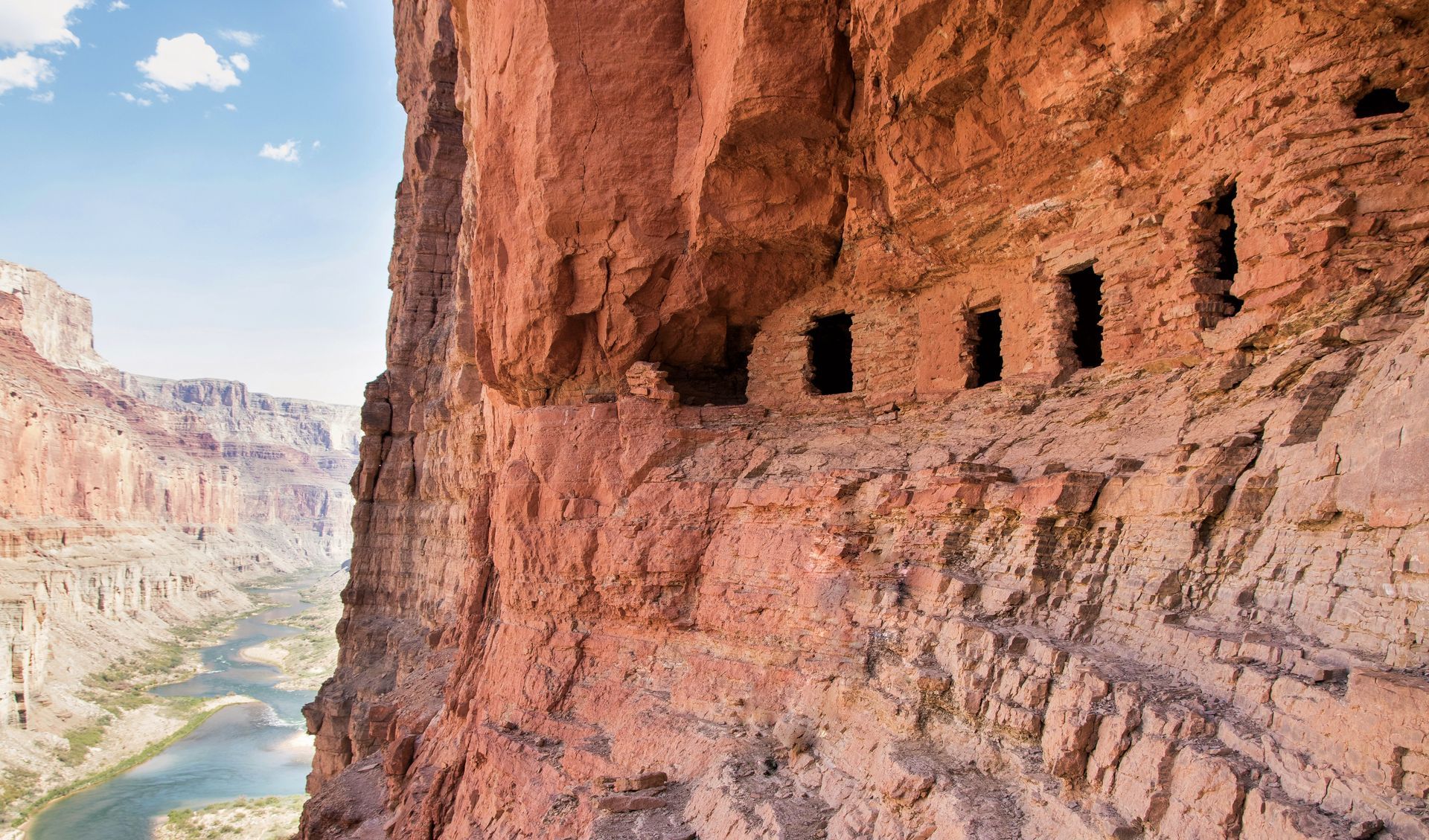 Red rock cliff face with ancient dwellings overlooking a river canyon.
