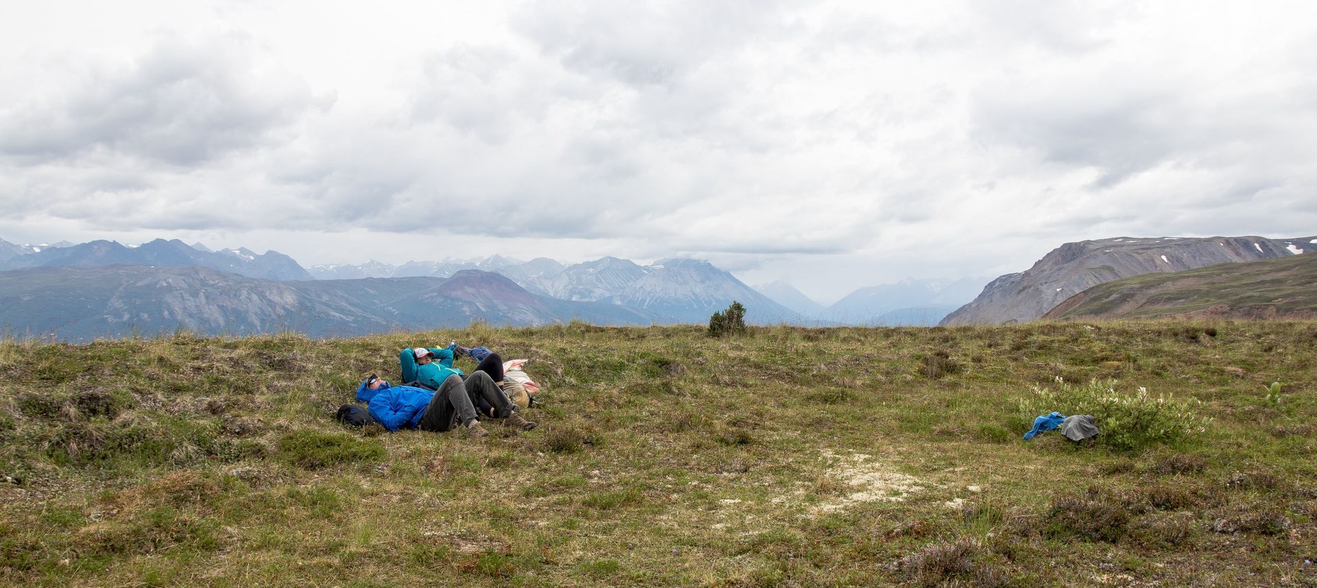 A mountain landscape with clouds overhead. Backpacks sit on the grassy ground, with mountains in the background.