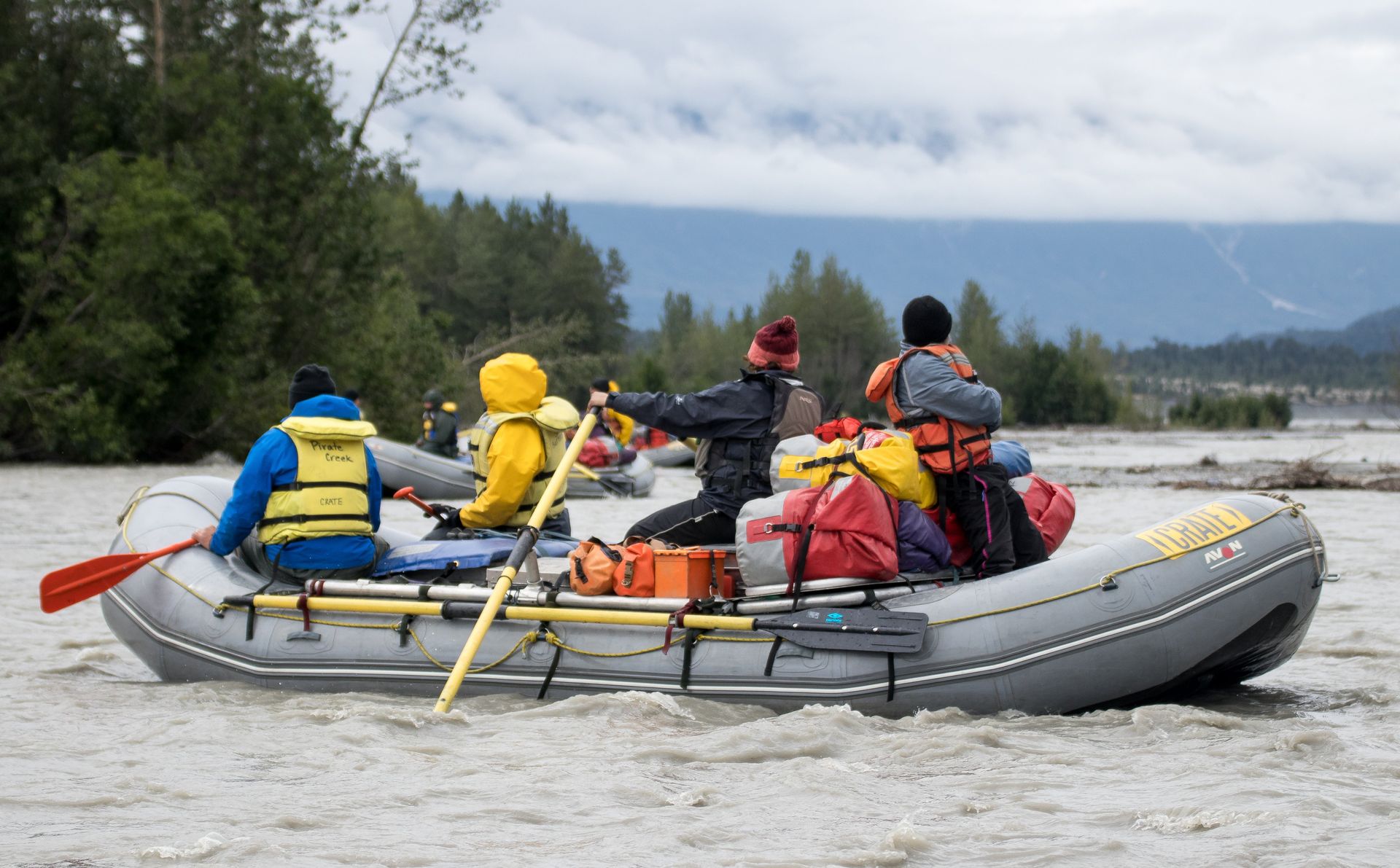 Rafting on a cloudy river. People paddle a gray raft with gear, wearing life vests and jackets. Forest in background.