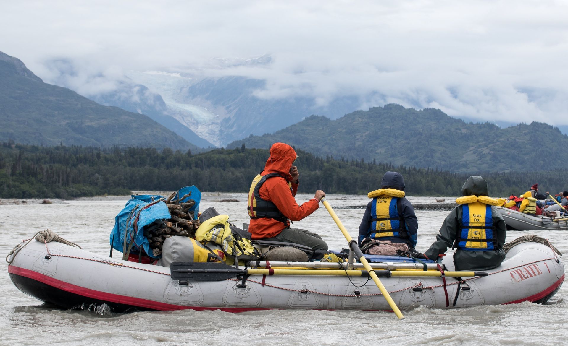 Rafters on a gray and white raft in a river, mountains in the background, overcast day.